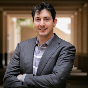 A smiling man with his arms crossed, wearing a gray blazer and gray and white striped button up