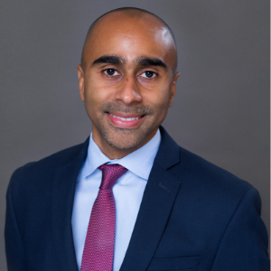 A smiling man wearing a navy blazer, light blue button up and pink tie