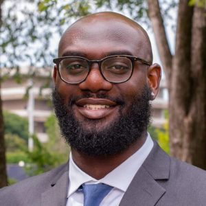 A smiling Black man wearing glasses, a gray blazer, white button up and blue tie