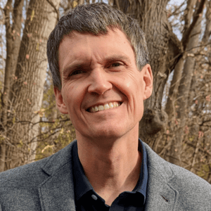 Smiling man in front of a tree, wearing a gray blazer with a dark blue button up