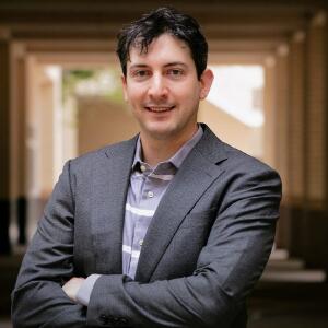 A smiling man with his arms crossed, wearing a gray blazer and gray and white striped button up