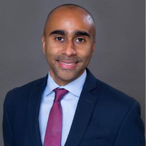 A smiling man wearing a navy blazer, light blue button up and pink tie