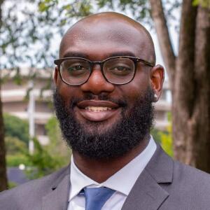 A smiling Black man wearing glasses, a gray blazer, white button up and blue tie