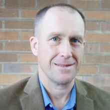 A smiling man wearing a brown blazer and a blue button up